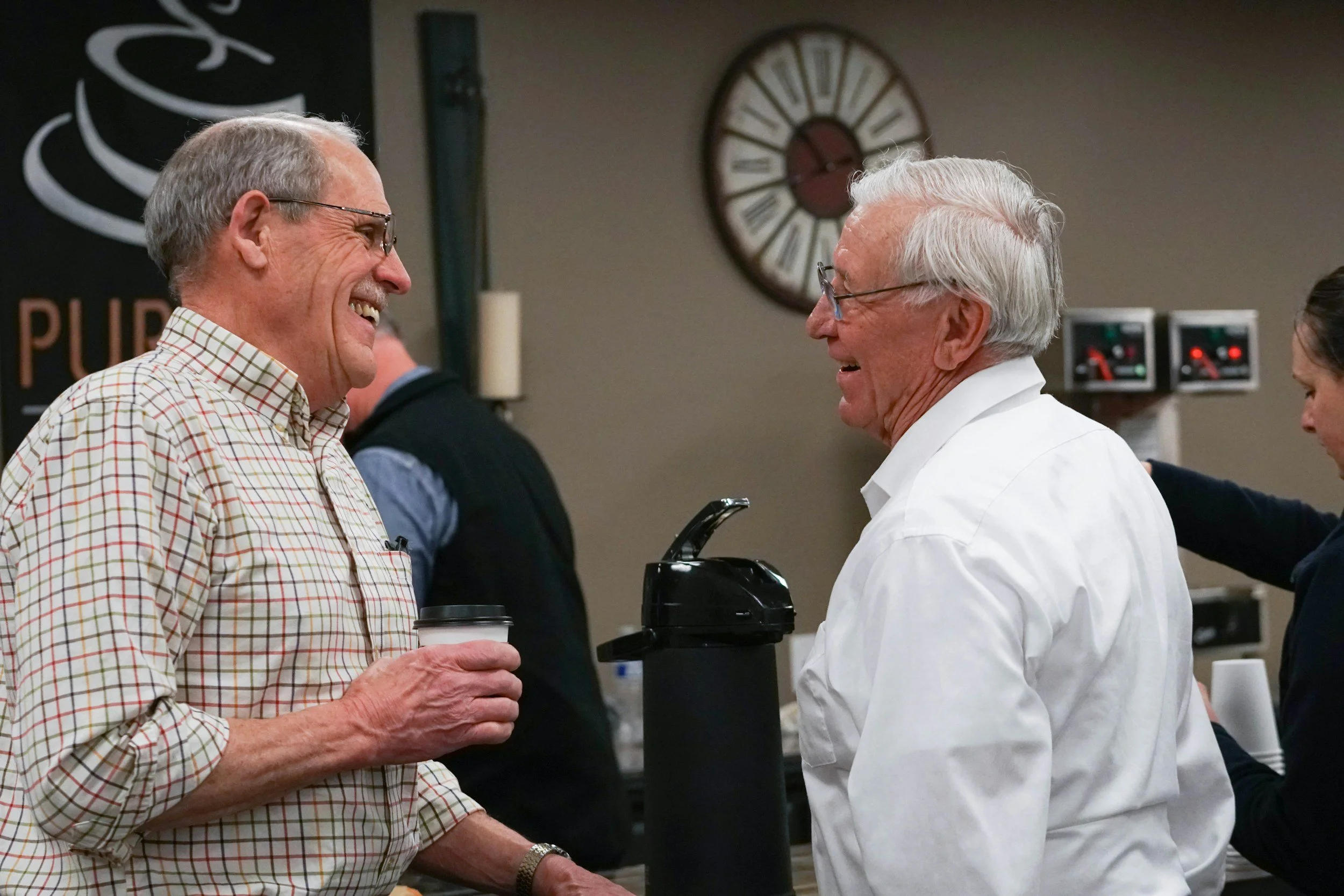 Two older men with glasses and gray hair are smiling and talking to each other in a kitchen or casual setting, holding coffee cups. One man wears a plaid shirt, and the other wears a white shirt.