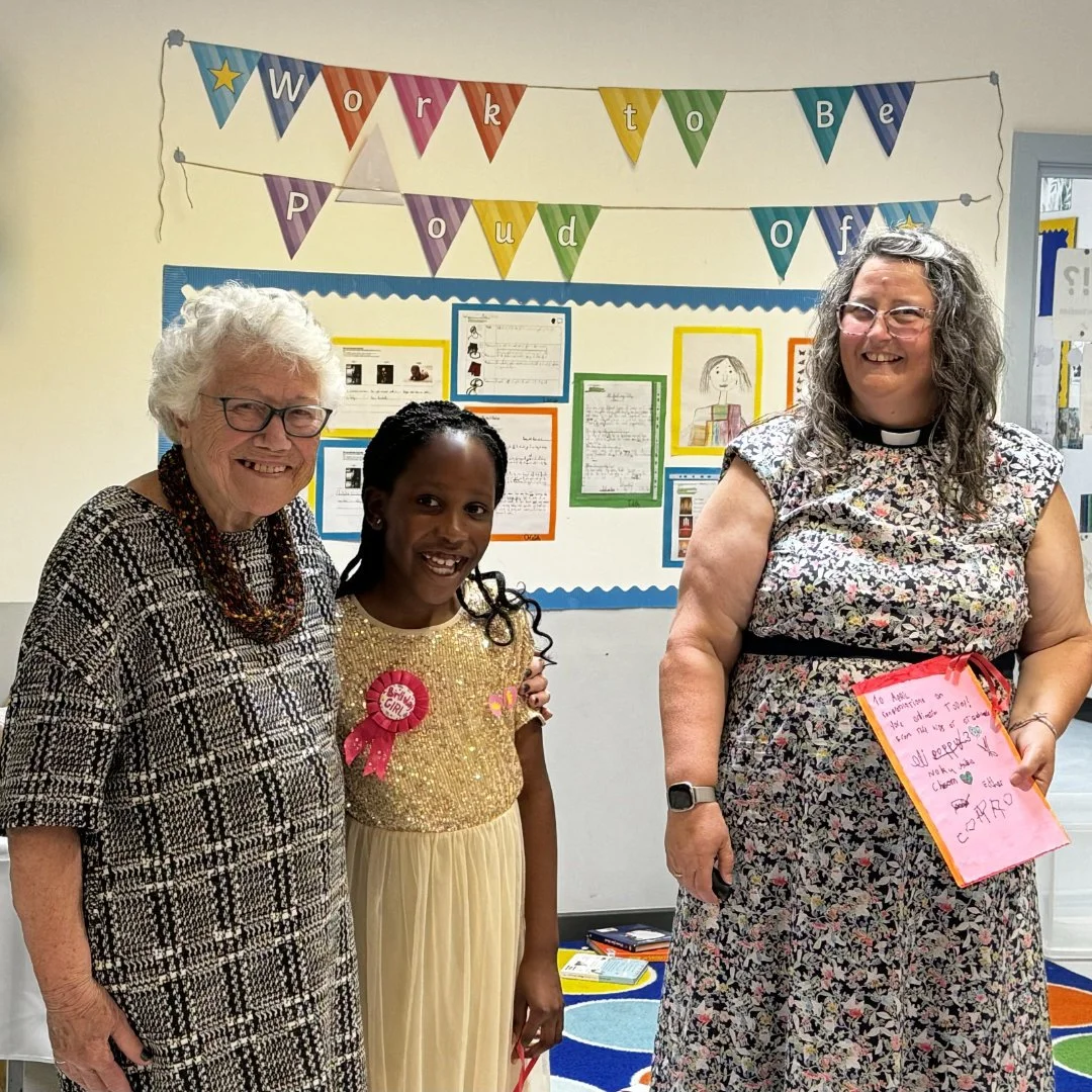 Three women standing together in a classroom, smiling. Behind them is a bulletin board with colorful papers and drawings, alongside a banner that reads 'Work to Be Proud of.' One woman is holding a pink paper with handwritten notes.