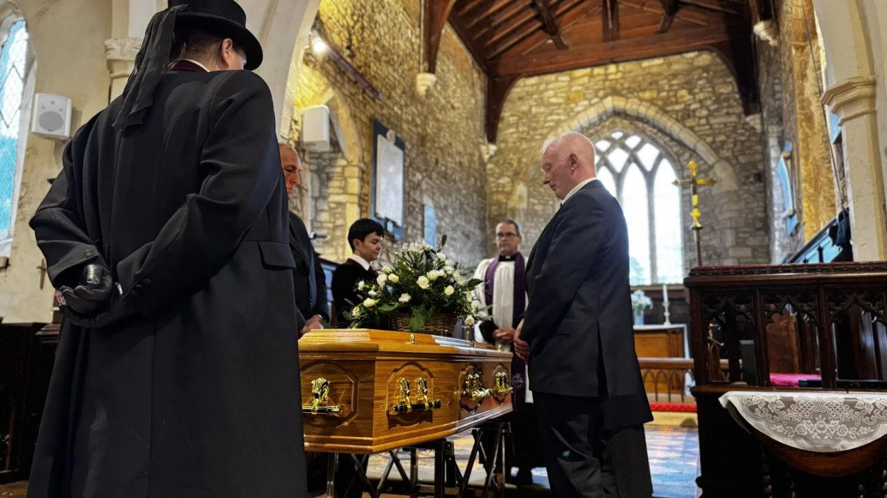 People mourning at a funeral service inside a church with stone walls, wooden beams, stained glass windows, and an altar in the background. A wooden coffin with gold handles is at the center, topped with a floral arrangement. One person is wearing a 