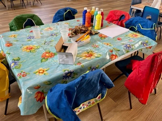 A table covered with a floral tablecloth in a room, set up for a craft or paint activity with cups, bottles of paint, brushes, paper, and other supplies, surrounded by chairs with jackets draped over them.