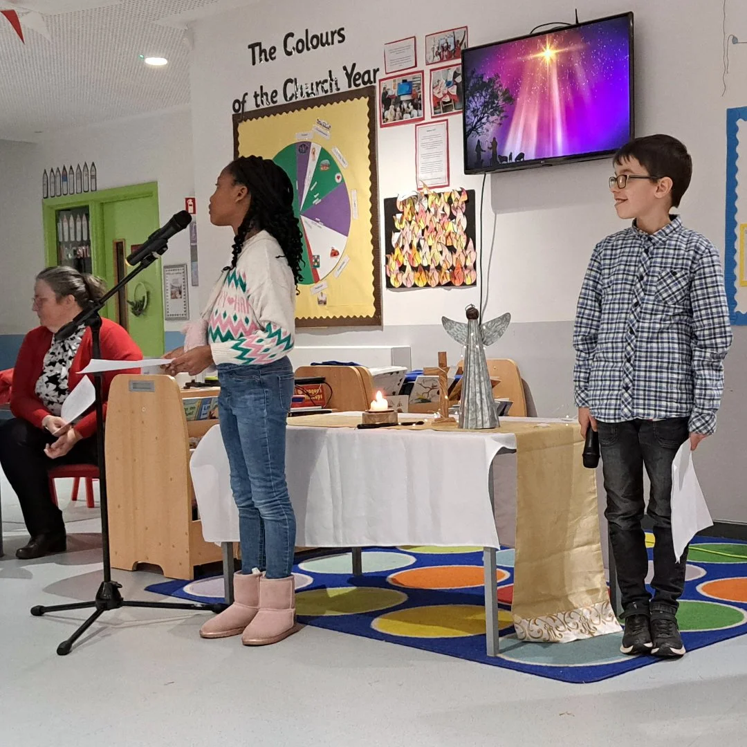 A school presentation with two children standing at microphones, one girl speaking and one boy waiting, in a classroom with teachers and colorful decorations, including a chart titled 'The Colours of the Church Year' and a television screen displaying a celestial scene.