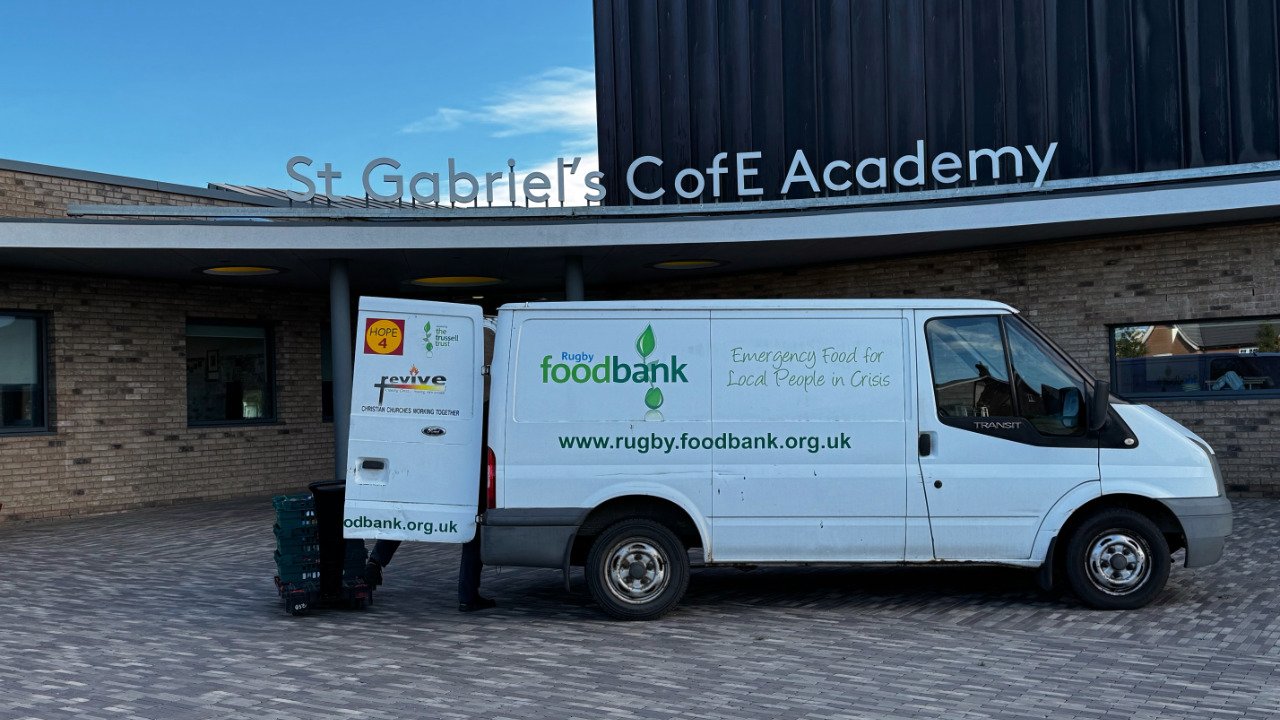 White van with 'Rugby Food Bank' logo and the message 'Emergency Food for Local People in Crisis,' parked in front of St. Gabriel's CofE Academy building.