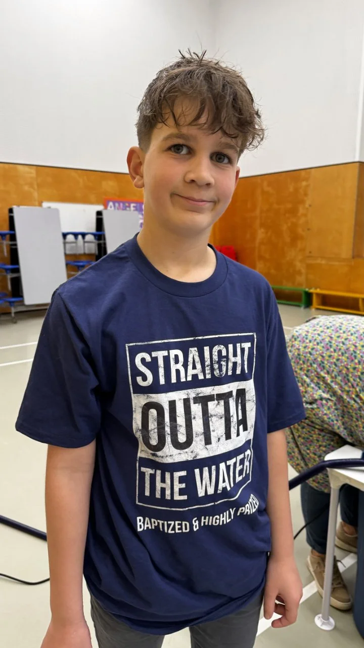 A young boy with curly hair and a slight smile, wearing a navy blue t-shirt that says 'Straight Outta The Water' and 'Baptized & Highly Prayed,' standing indoors in a room with gym mats and chairs.