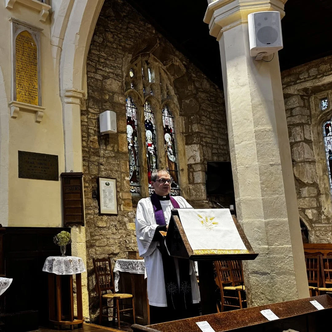 A priest standing at a lectern inside a stone church, with stained glass windows and religious plaques on the wall behind.