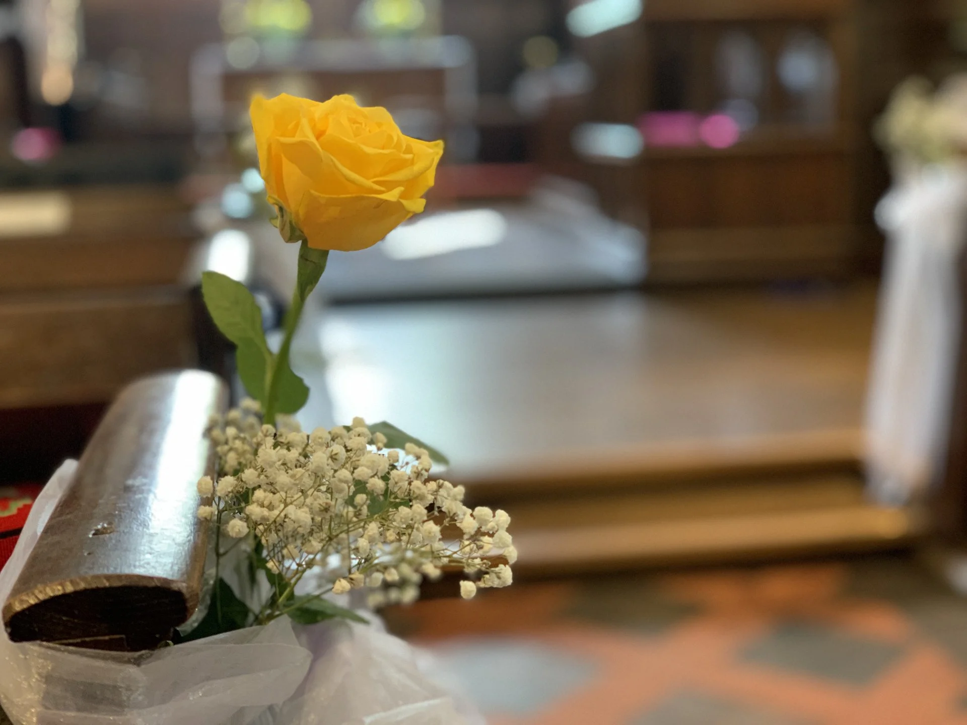A single yellow rose with green leaves in a small bouquet, placed in a white wrapping, sitting on a wooden pew inside a church or chapel with blurred background.