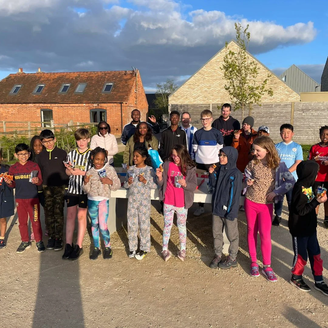 A group of children and adults gathered outdoors, standing on a gravel surface with houses and a wooden fence in the background, enjoying snacks.