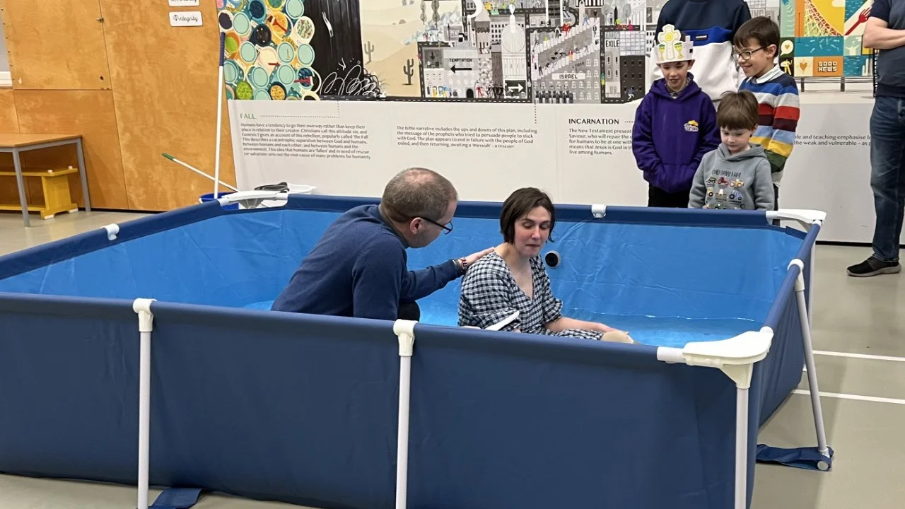 A woman is inside a small blue baptism pool, with two men beside her, one reaching over her shoulder. Four children and one adult are standing outside watching.