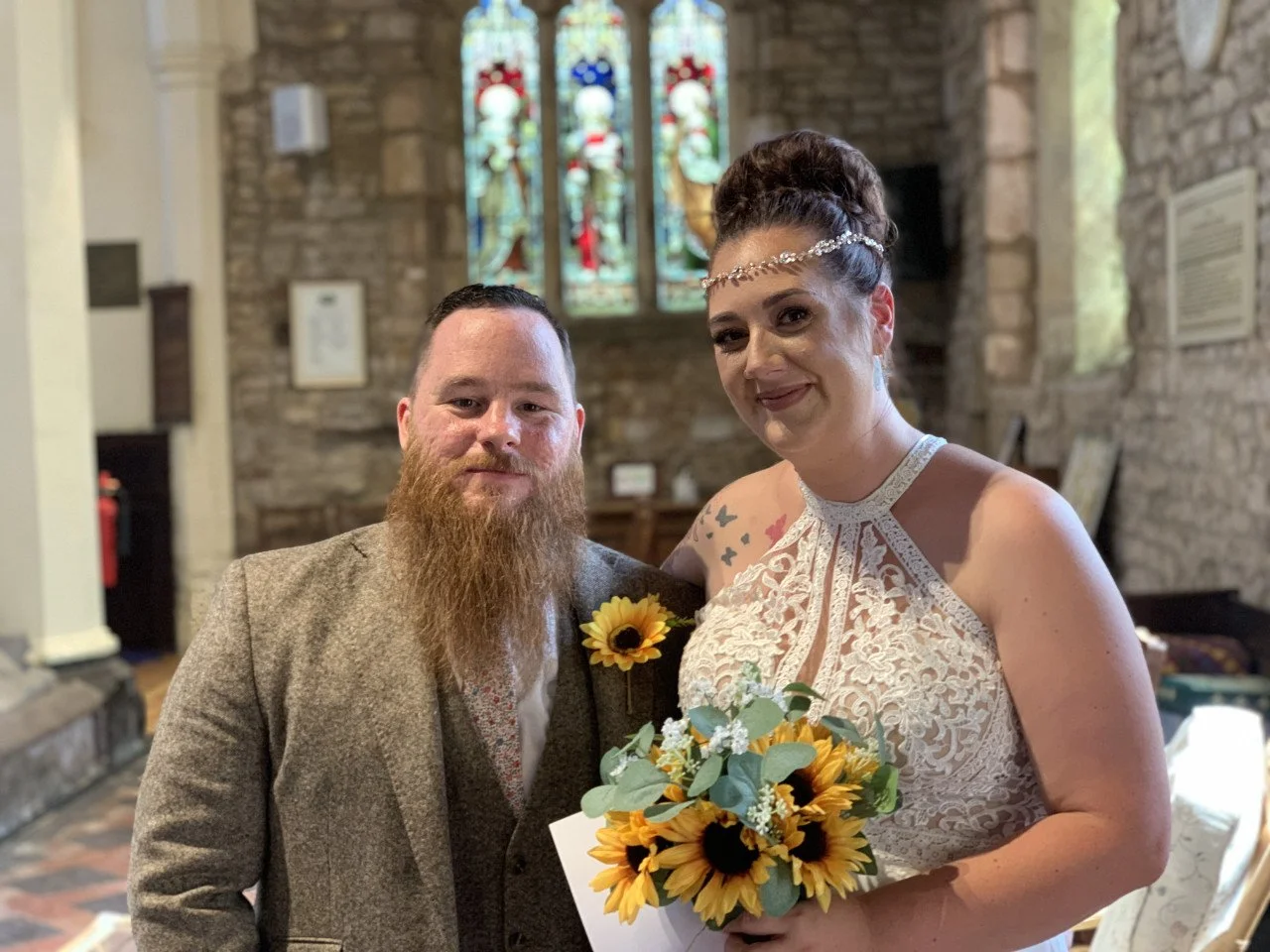 A bride and groom pose inside a church, with stained glass windows and stone walls in the background. The bride holds a bouquet of sunflowers and white flowers, wearing a lace wedding dress, and the groom has a beard and is dressed in a brown suit ja