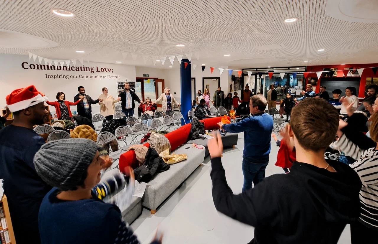 People celebrating and dancing in a community center decorated with festive bunting, some wearing holiday accessories, during a holiday gathering.