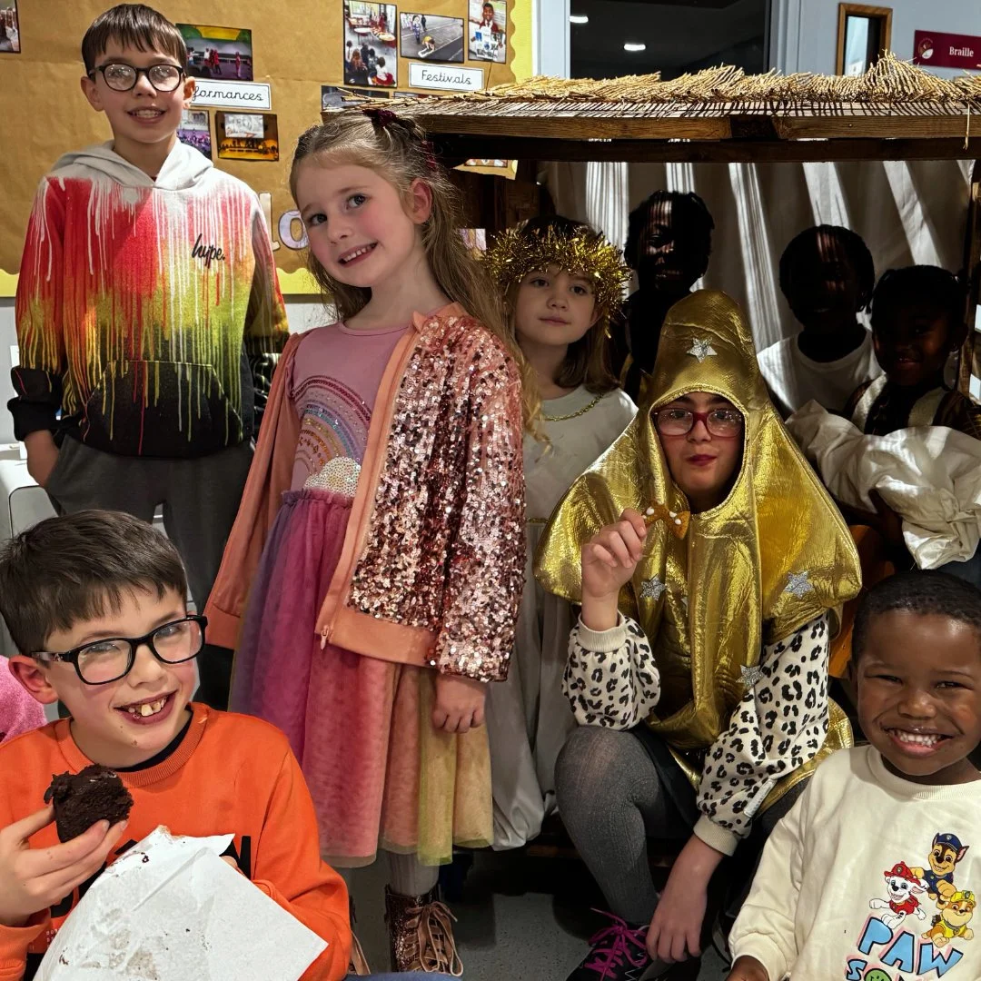 Children dressed in costumes for a school play, including angels, a wise person with a gold cape, and others, posing together inside a classroom.