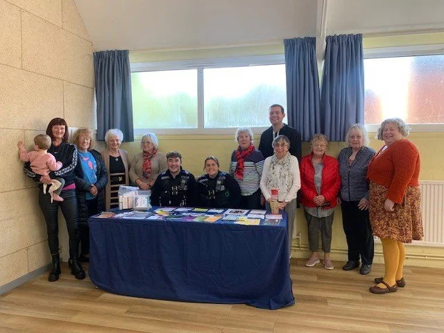 Group of elderly women and two police officers standing behind a table with papers and pamphlets in a room with windows, blue curtains, and wooden floors.