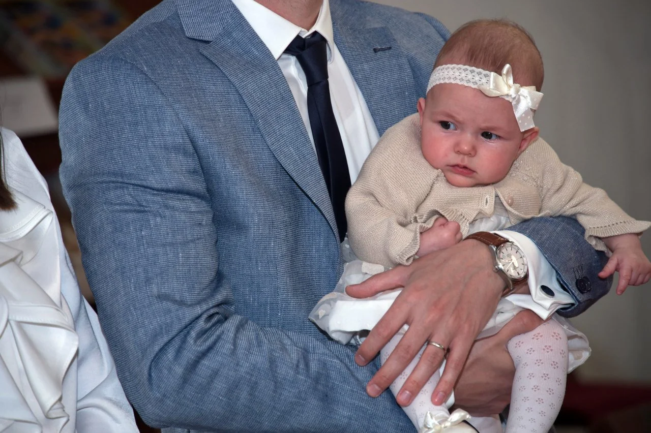 A man in a blue suit holding a baby girl with a white headband and cream-colored cardigan at a christening event.