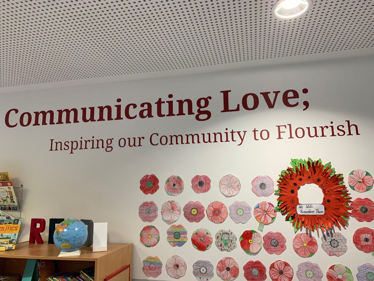 A wall with the phrase 'Communicating Love; Inspiring our Community to Flourish' in large red letters, decorated with colorful paper flowers and a wreath with a sign that says 'We Will Remember Them.' There is a bookshelf with books and a globe on top.