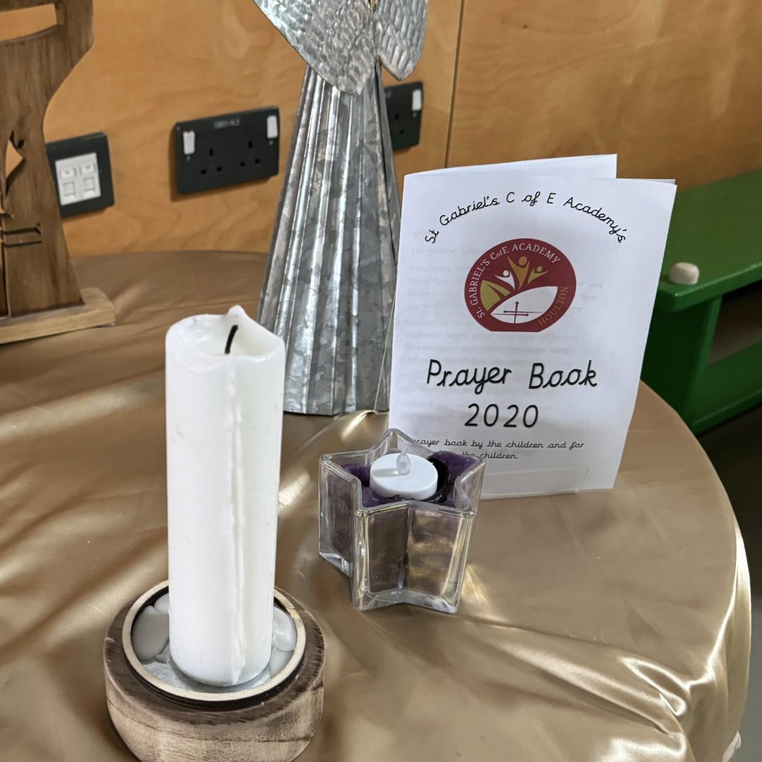 A prayer book from St. Gabriel's C of E Academy 2020, a lit prayer candle, a decorative silver angel sculpture, and a small candle in a glass holder on a gold-colored table.
