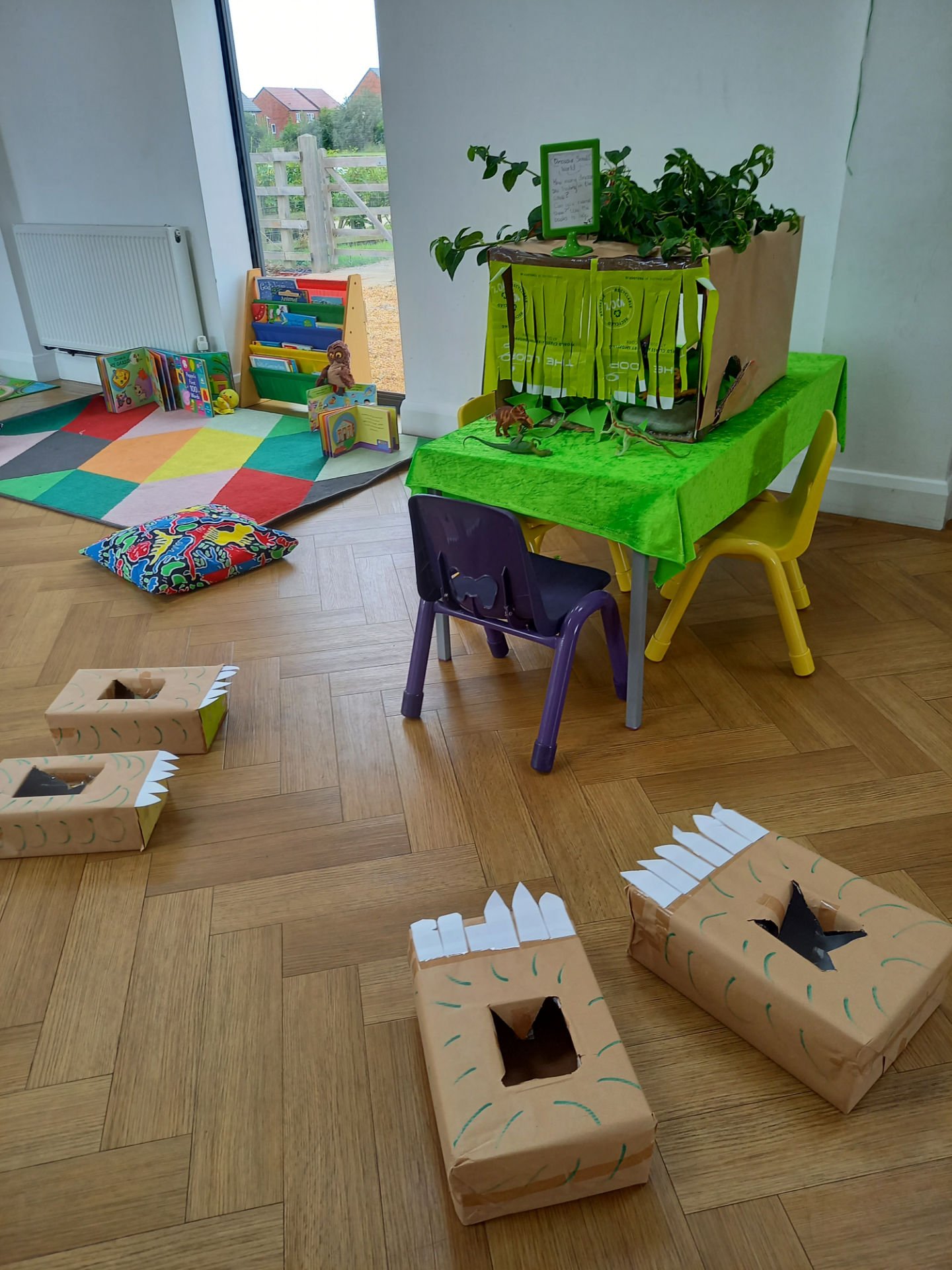 A room with a wooden floor, decorated with paper boxes shaped like dinosaurs, a small table with colorful chairs, a bookshelf with children's books, and a window showing a garden and house outside.