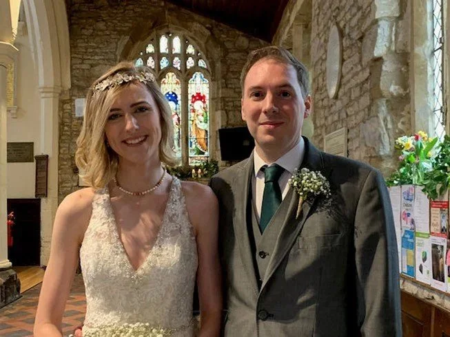 A bride and groom standing inside a stone church with stained glass windows and floral decorations, smiling at the camera.