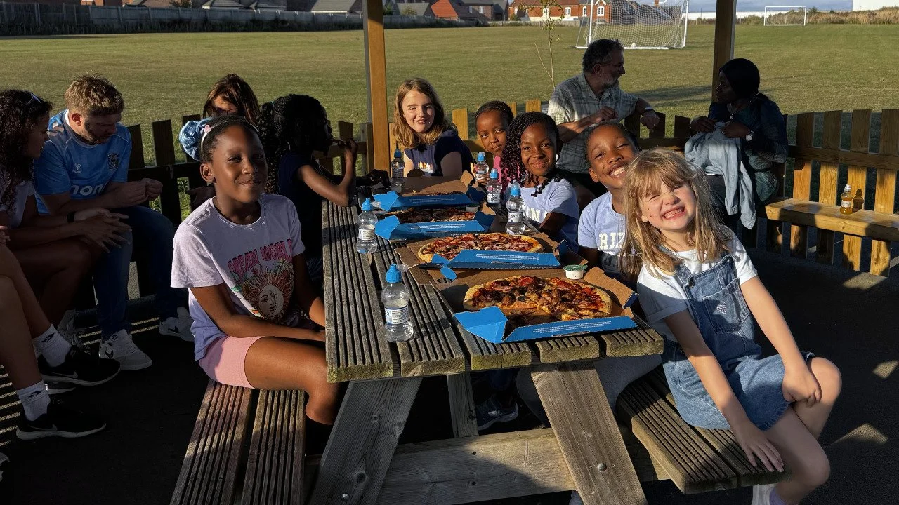 Children sitting at a picnic table with pizza boxes, some with pizza slices, in a park or playground, with a grassy field and goalpost in the background.