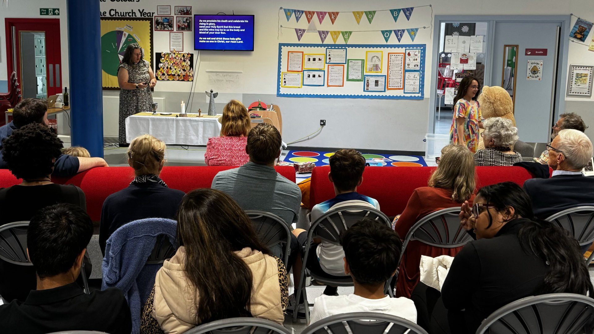 A group of people attending an indoor presentation in a church or community hall. A woman stands at the front near a table, speaking to the audience, with a large screen displaying text behind her. The audience is seated, facing the presenter, with some people in the foreground and others visible on the sides. The walls are decorated with colorful posters, banners, and bulletin boards.