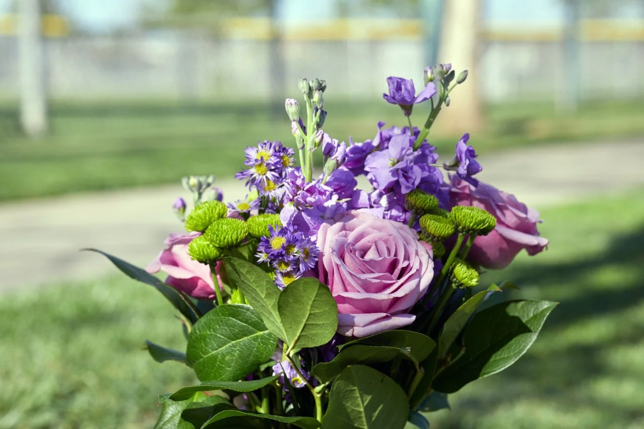 A bouquet of pink roses, purple lisianthus, small purple and yellow daisies, and green foliage outdoors with a blurry background.