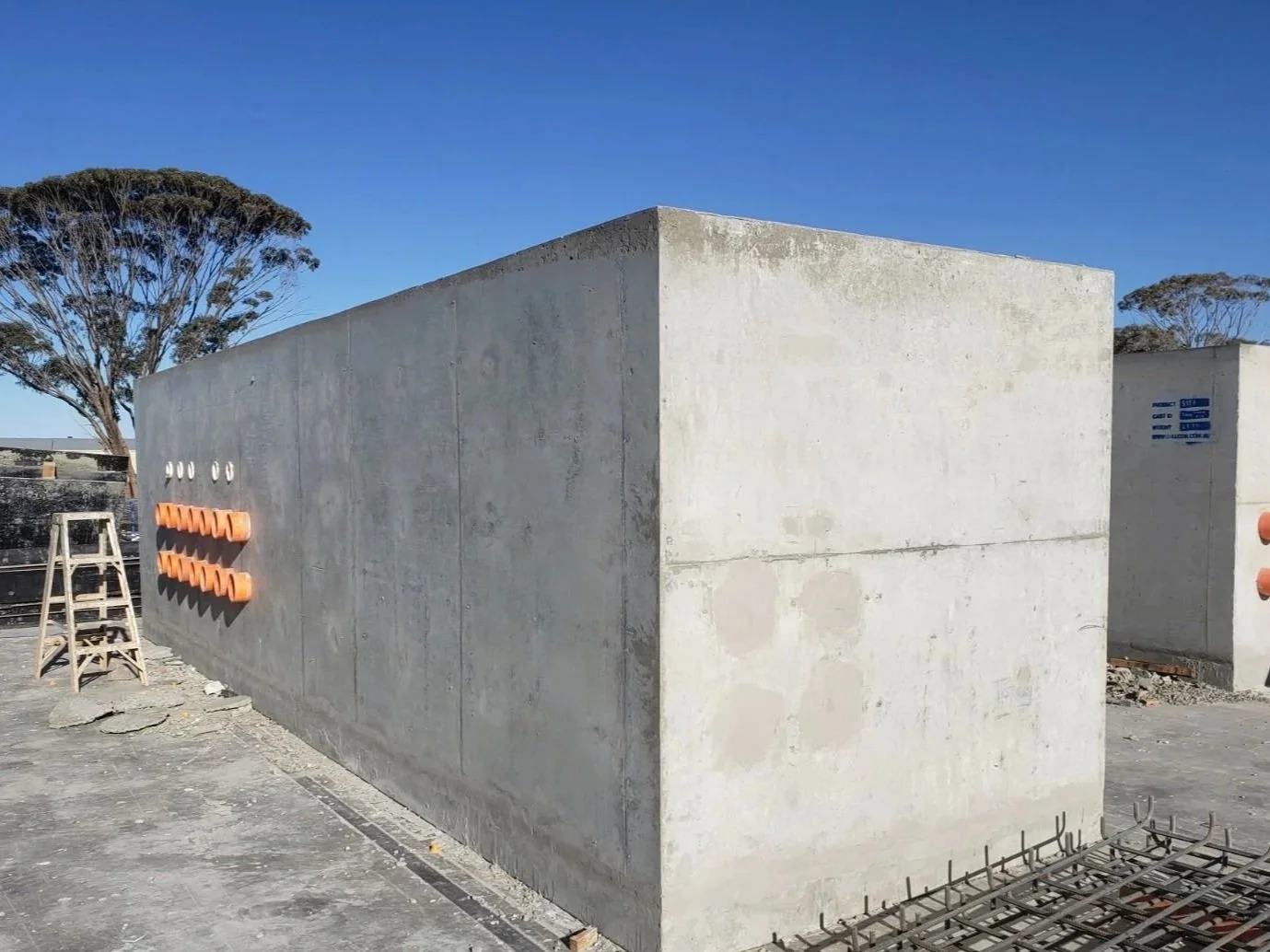 Construction site with a large concrete wall, orange and white pipe fixtures, a wooden ladder, and tree in the background under a clear blue sky.
