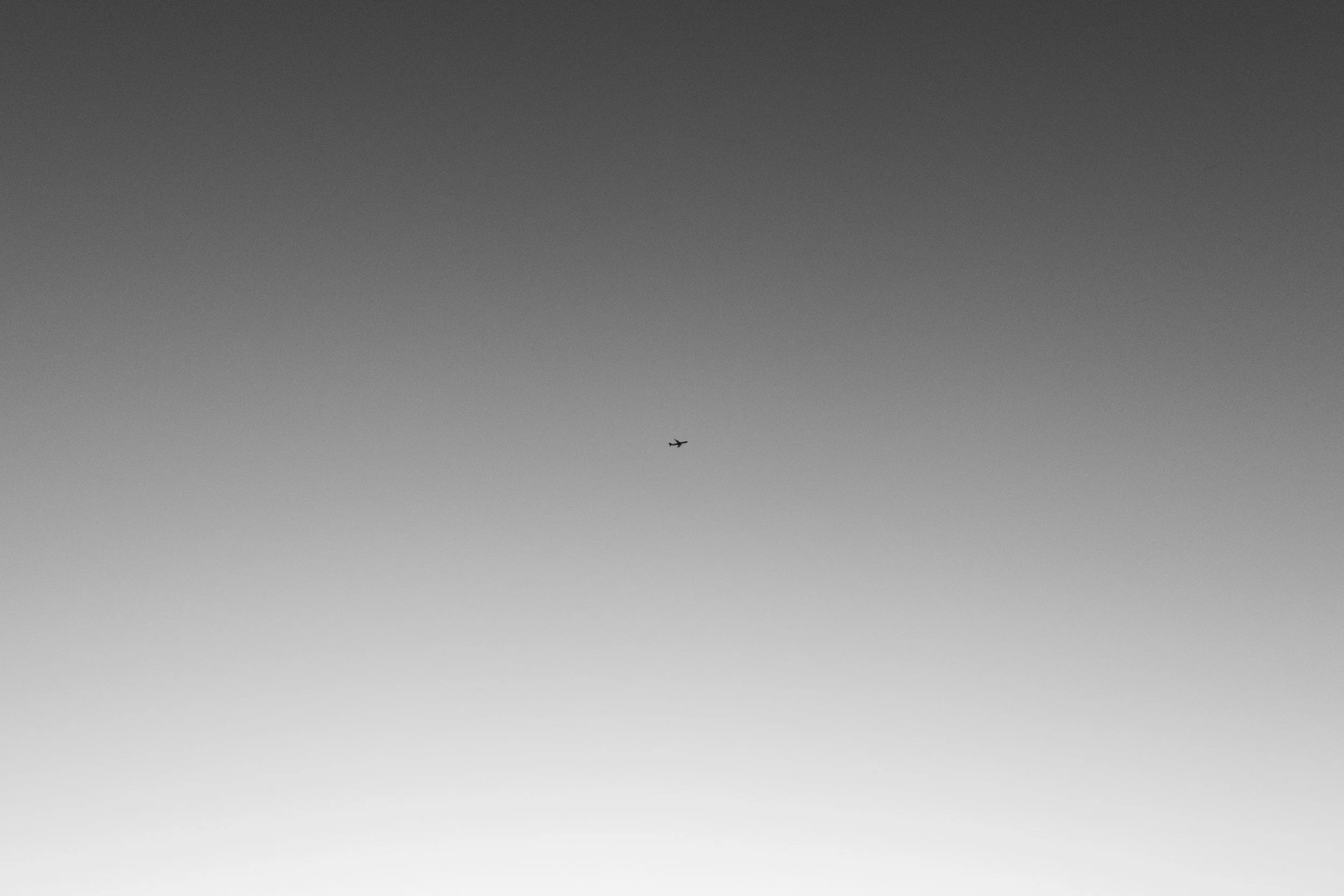 Black and white photo of a small airplane flying in a gradient sky from dark to light.