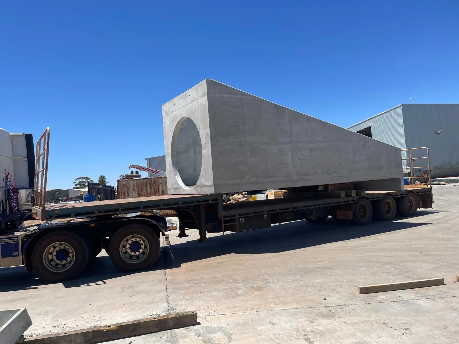 A large, rectangular concrete structure with a circular hole on one side is loaded onto a flatbed truck at an industrial site. The sky is clear and blue.