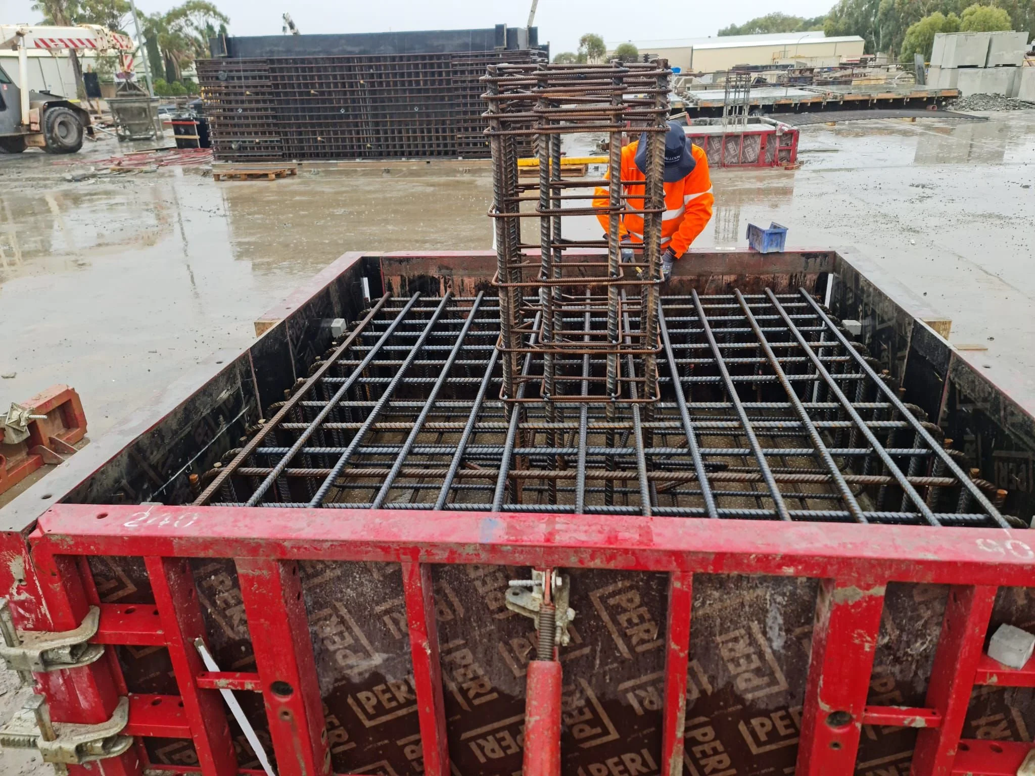 Construction site with rebar framework and a worker in an orange jacket and blue cap installing rebar on a concrete foundation.