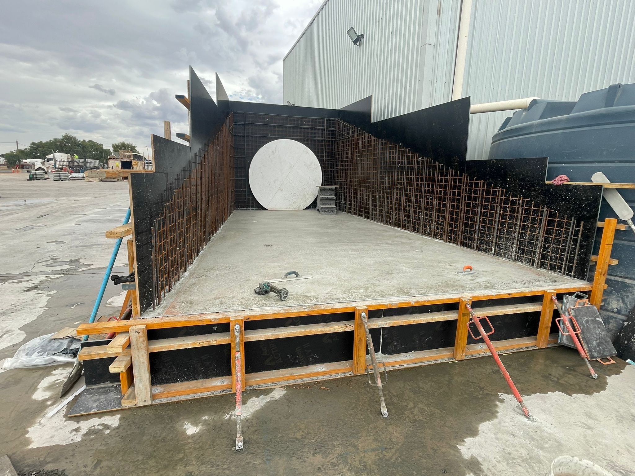 Construction site with a concrete slab foundation and formwork for a small raised platform, with steel reinforcement and black panels, next to a large industrial building and a water tank.