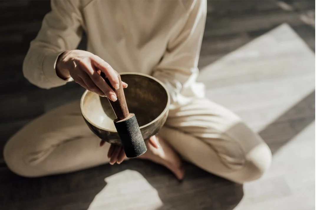 Person sitting cross-legged on wooden floor, holding a singing bowl and a mallet.