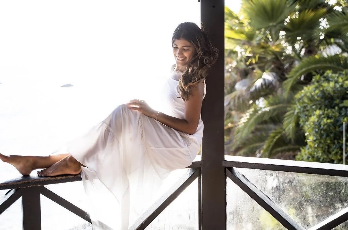 Young woman in a white dress sitting on a wooden balcony railing, smiling and looking down, tropical trees in the background
