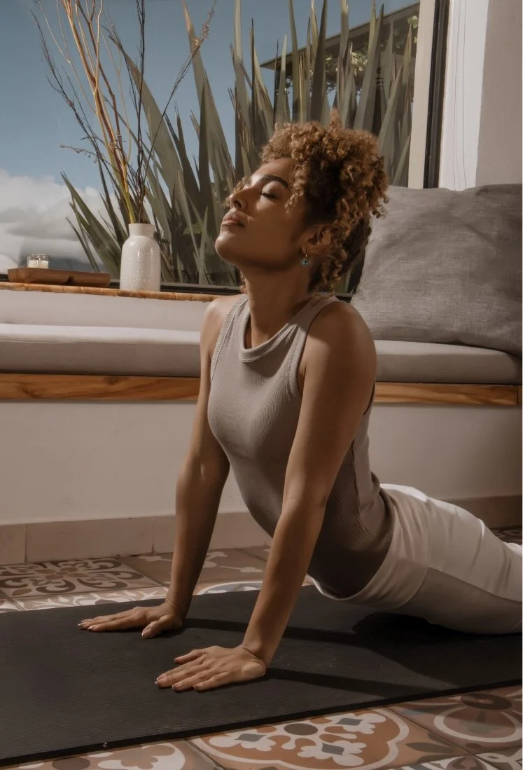 A woman practicing yoga in a room with large windows, sitting on a black yoga mat, with plants visible outside the window, wearing a beige sleeveless top and light-colored pants, with her eyes closed and head tilted back.