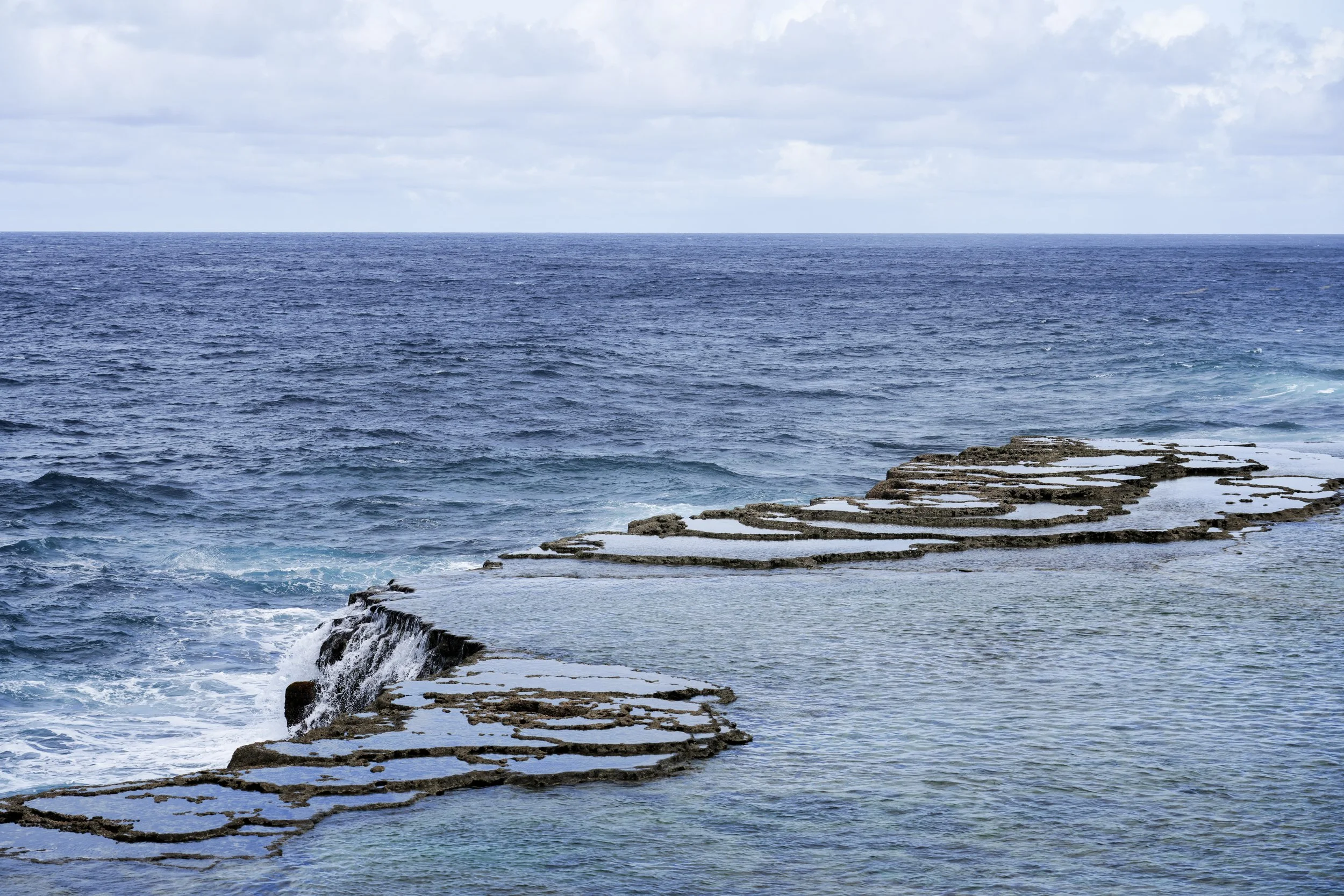 Tonga: Rock formations on the shoreline by the ocean with waves crashing against them under a cloudy sky.