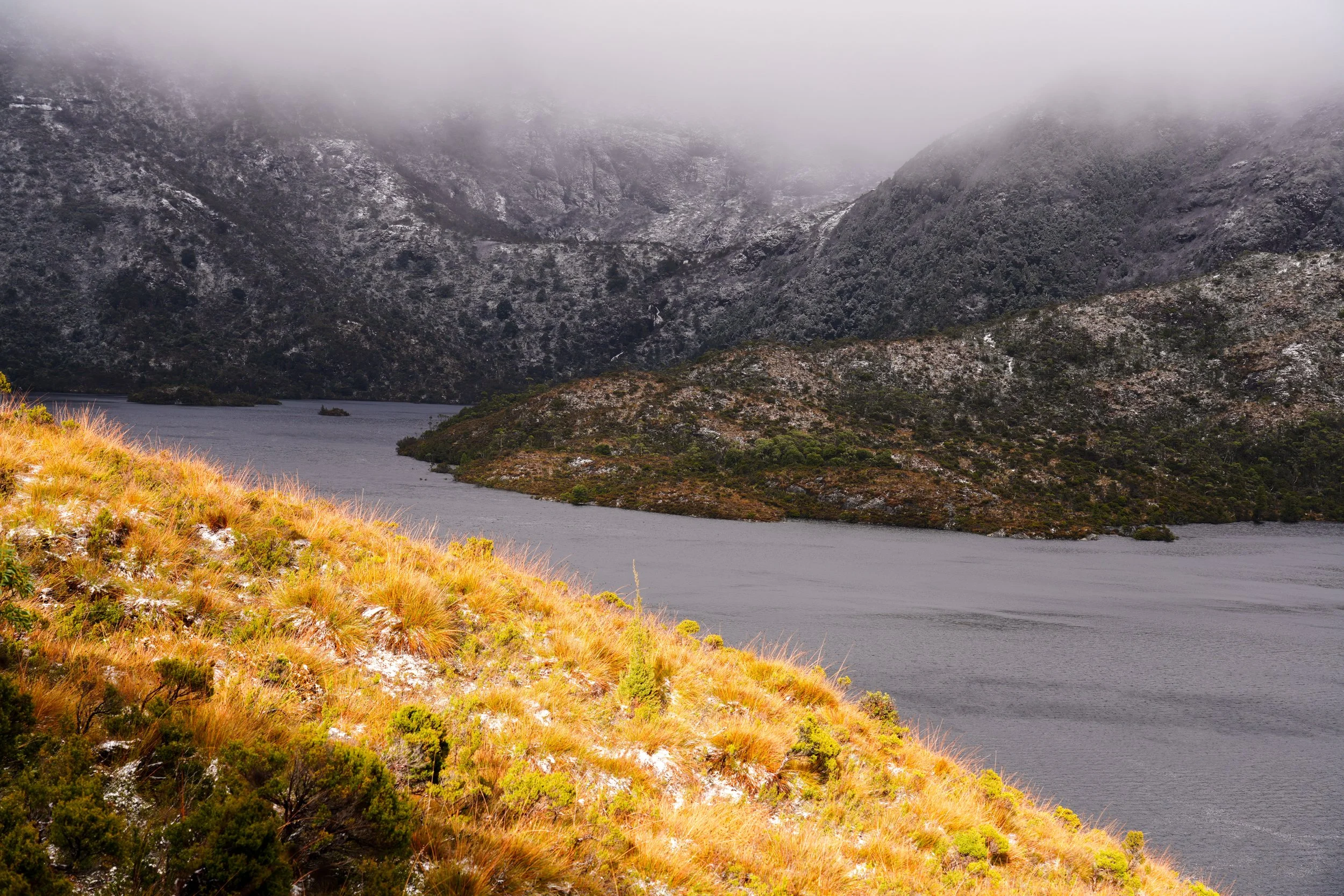 Cradle Mountain, Tasmania: A landscape of a lake surrounded by mountainous terrain with mist covering the mountain peaks and dry, grassy land in the foreground.