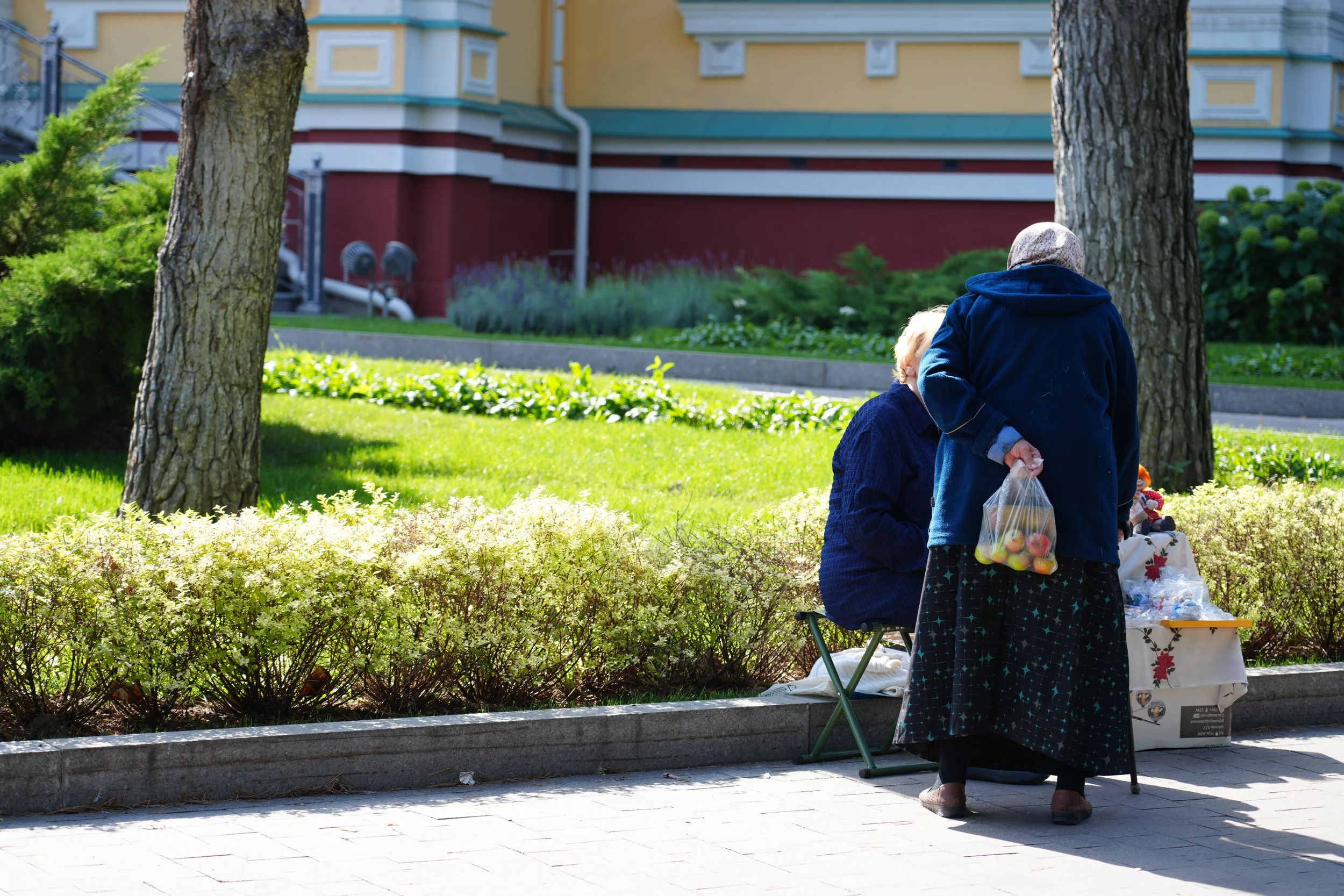 Almaty, Kazakhstan: Two women, one sitting on a small stool, the other standing, exchanging items at an outdoor market stall in a park, with green grass, shrubs, and trees in the background.