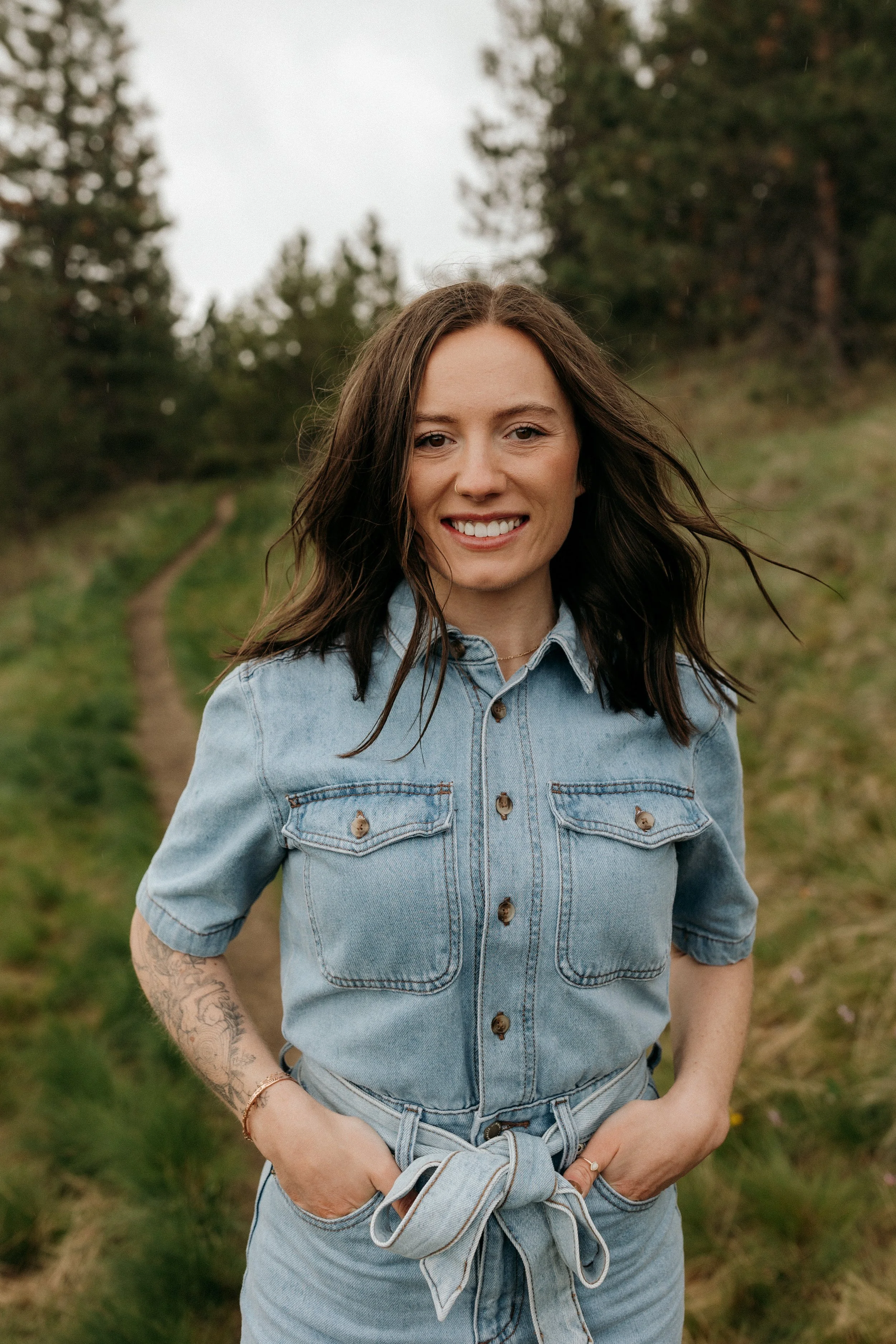 A young woman standing outdoors on a trail in a grassy area with trees, smiling at the camera, wearing a denim shirt and jeans.