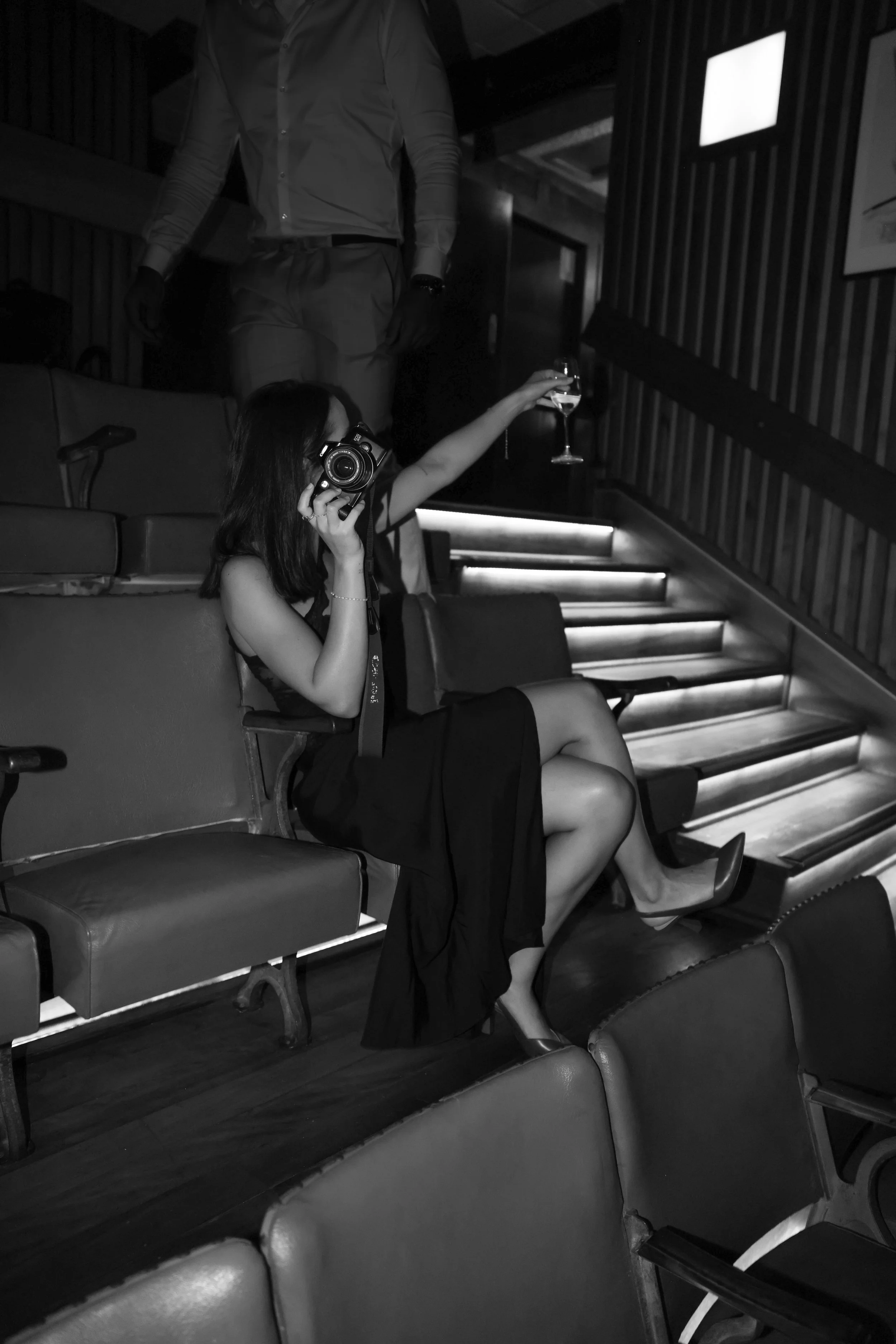 A woman sitting in a theater seat, wearing a black dress and high heels, holding a glass of wine, with her other hand near her face, and a person standing behind her, holding a camera.