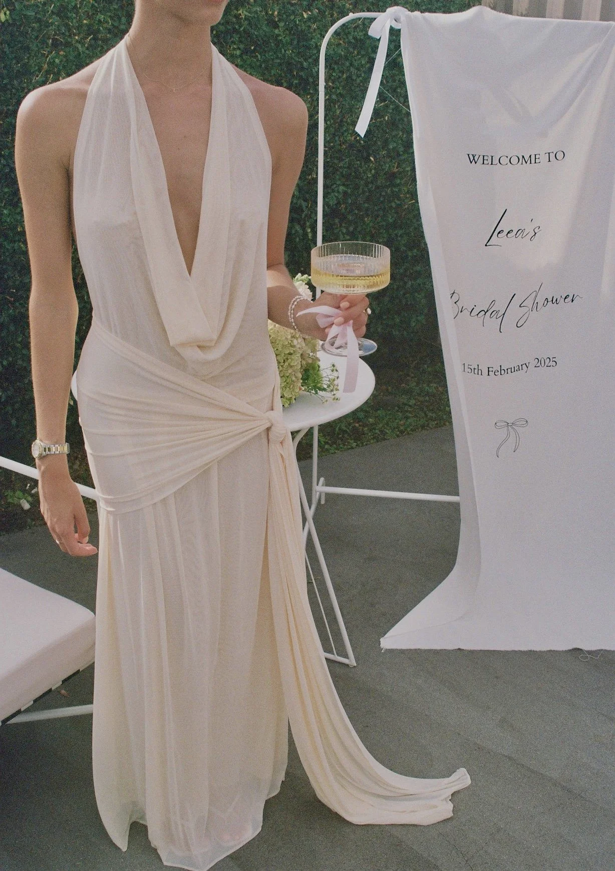 A woman in a cream-colored gown holding a cocktail glass at a bridal shower event. A welcome sign with event details is visible next to her.