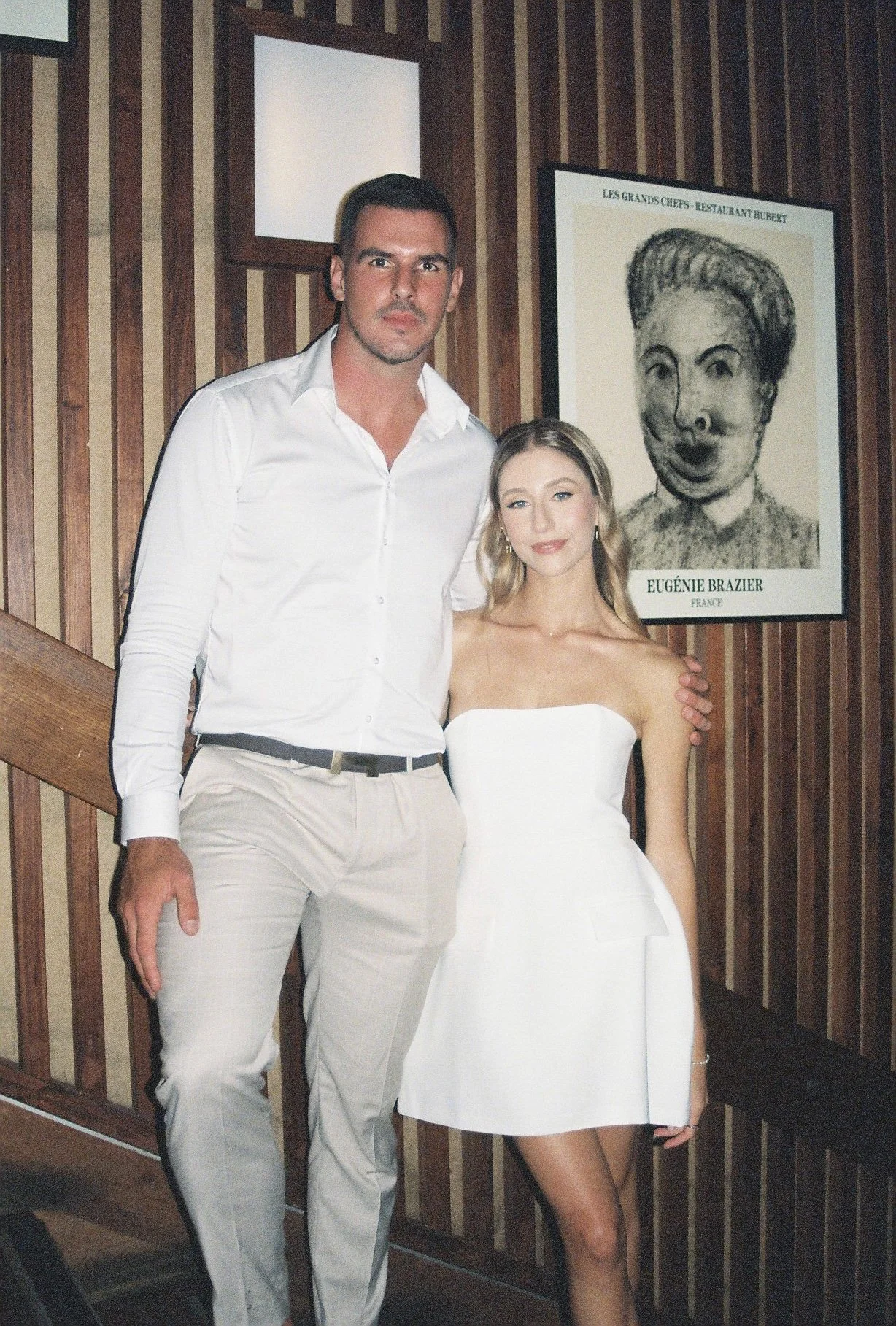 A man and woman posing together in front of a wooden wall and a framed art piece.