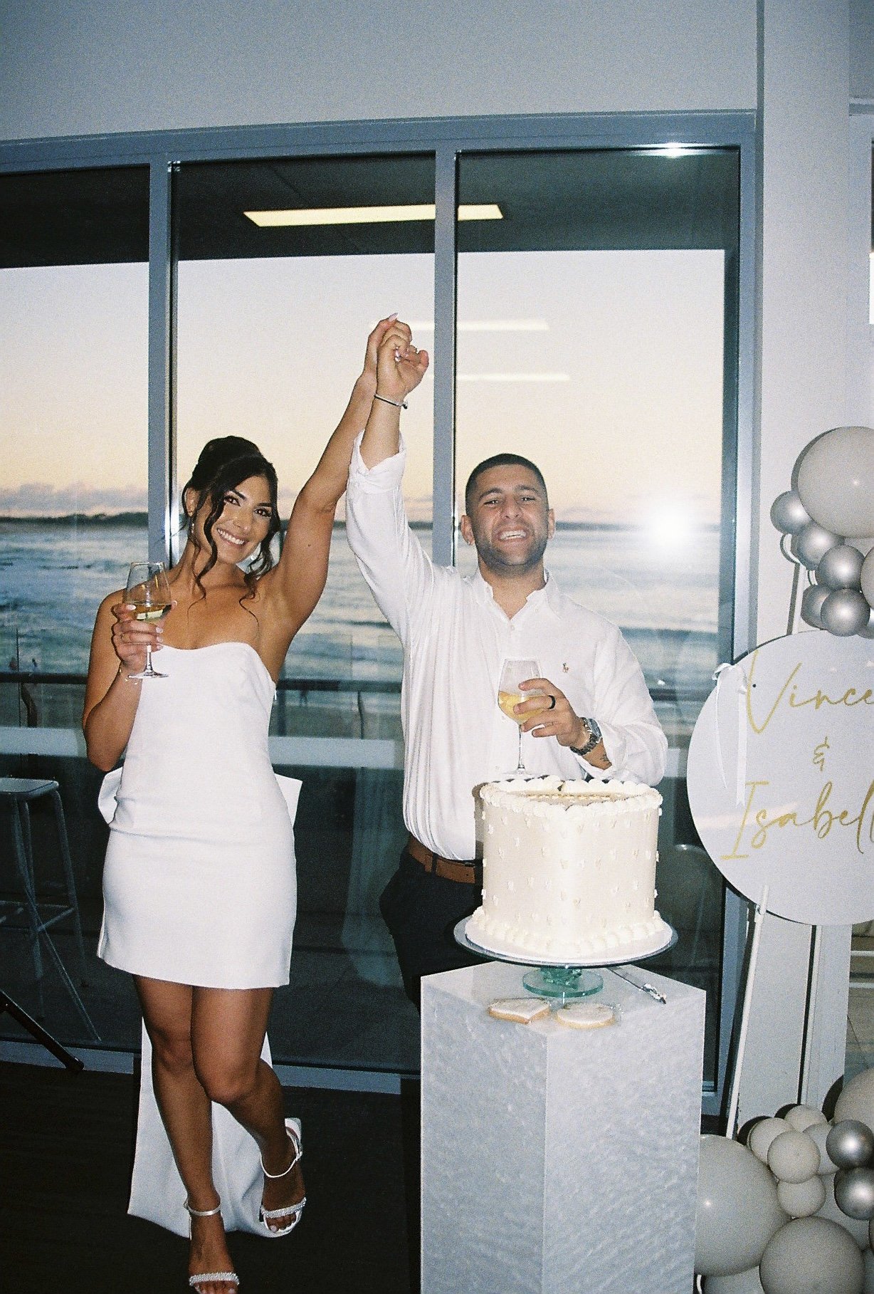 A woman and a man celebrating with glasses of white wine, standing next to a large white birthday cake at an indoor event by a window overlooking a body of water at sunset. The woman is wearing a white strapless dress, and the man is wearing a white 