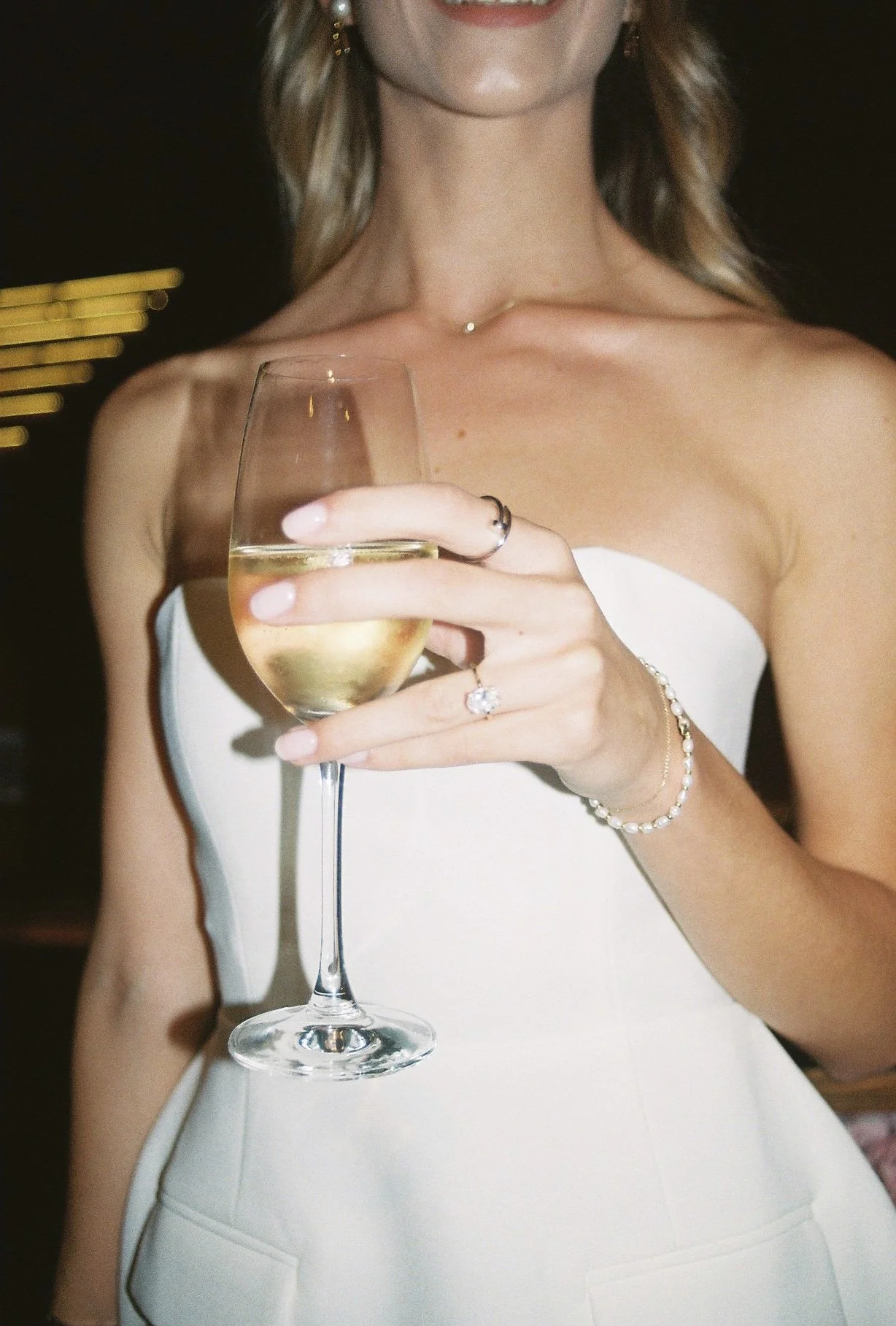 A woman wearing a strapless white dress holding a glass of white wine, with jewelry including rings, a bracelet, earrings, and a necklace, smiling in a dimly lit setting.