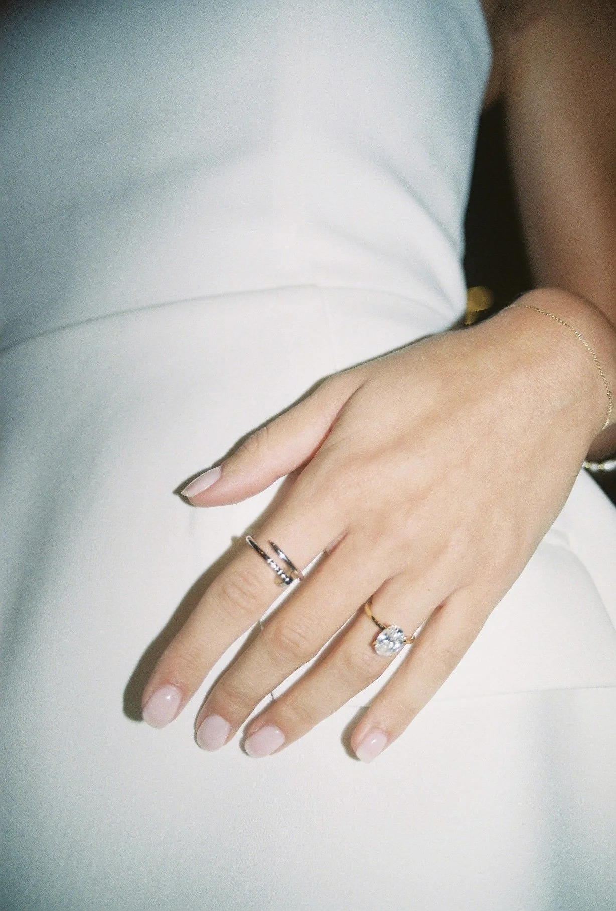 Close-up of a woman's hand resting on a white surface, wearing multiple rings and a delicate bracelet, with a white top visible in the background.