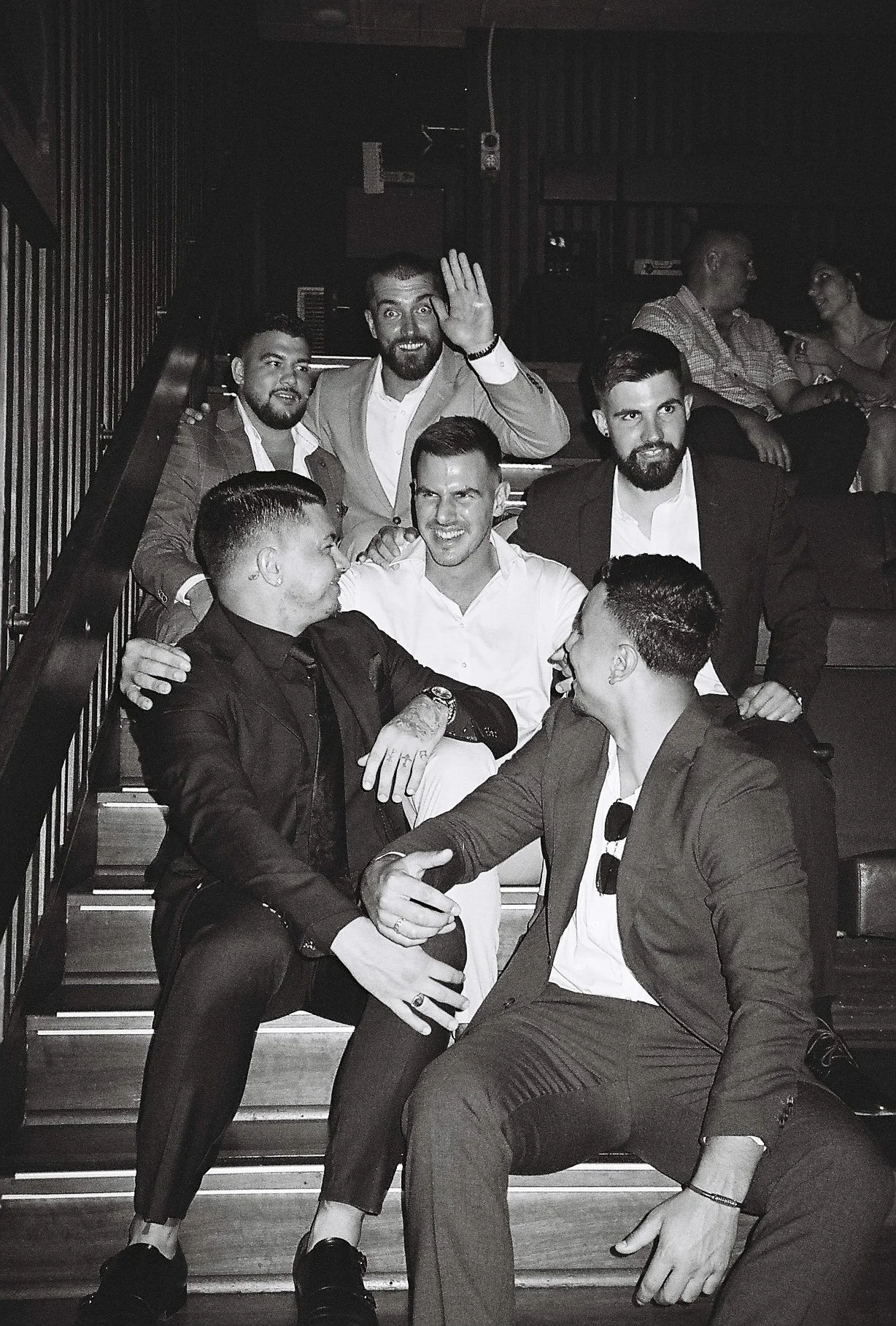 Group of men posing on stairs in a social setting, with some sitting and others standing, engaged in conversation and smiling.