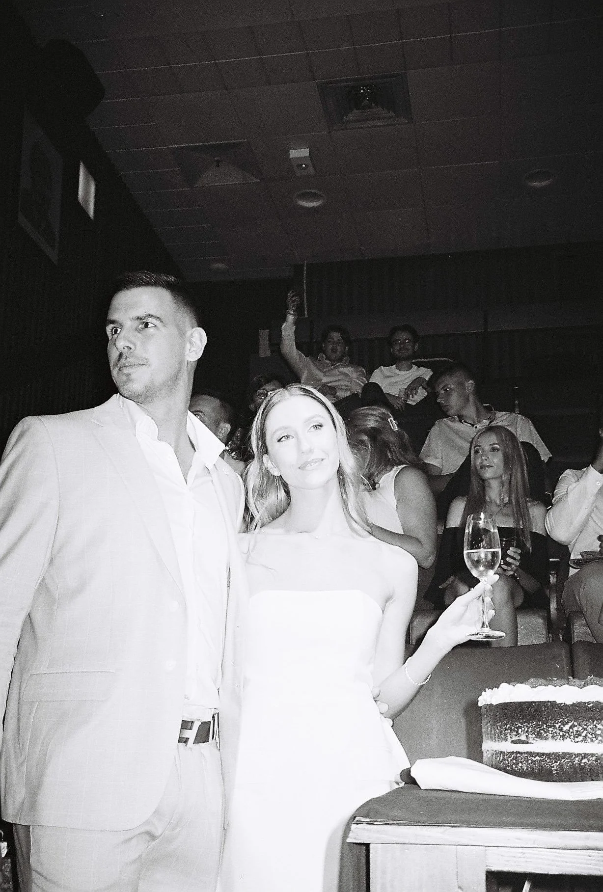 A group of people at a celebration or party, including a man in a light-colored suit and a woman in a white dress holding a glass of wine, with a cake in front of them. Others are seated in the background, some smiling and enjoying the event.
