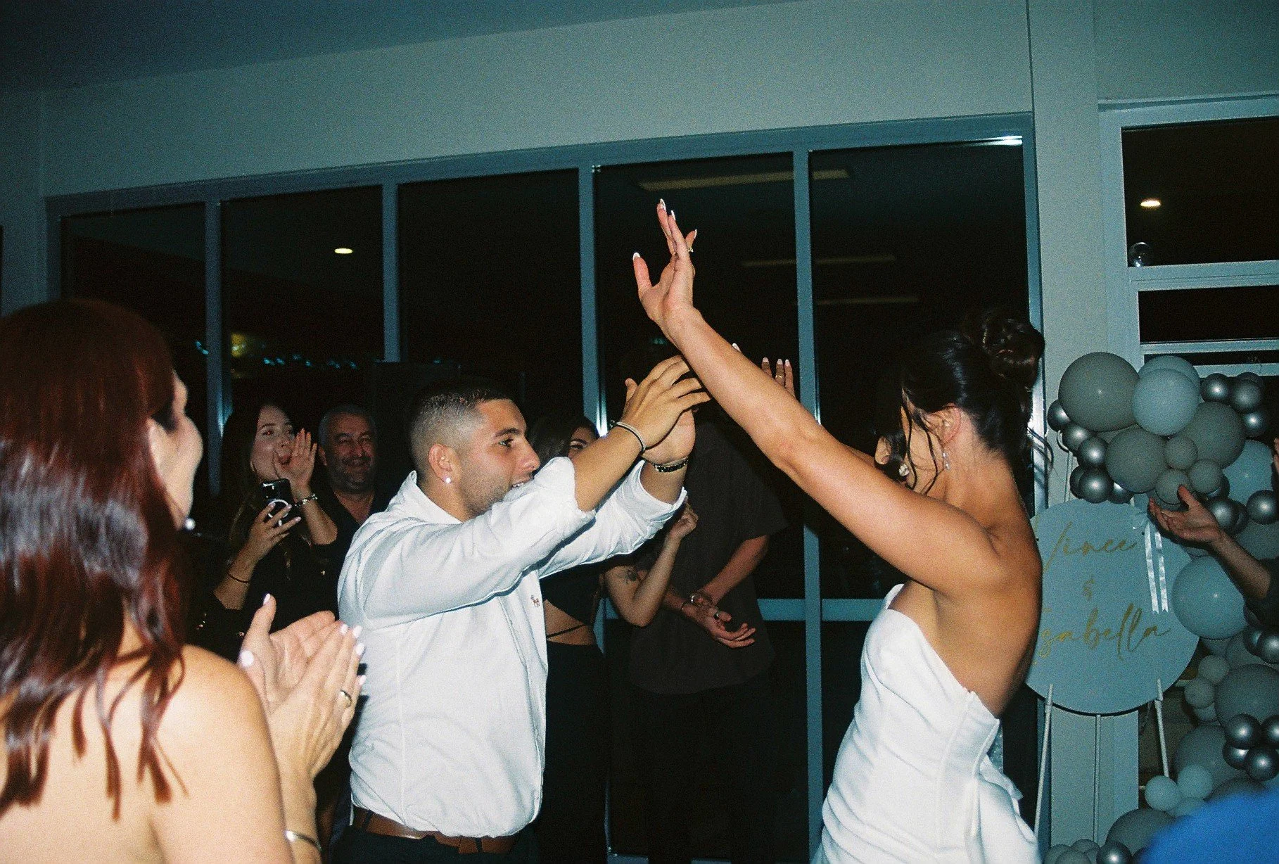 A woman in a white dress and man in a white shirt exchange a high-five during a celebration, surrounded by smiling guests in a decorated indoor space with balloons and a sign.