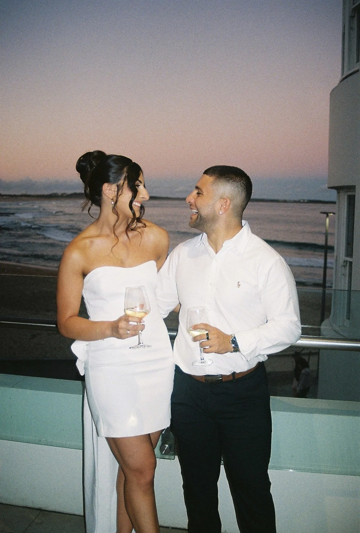 A couple enjoying a drink at sunset by the beach, smiling and looking at each other, with a glass of white wine in their hands.