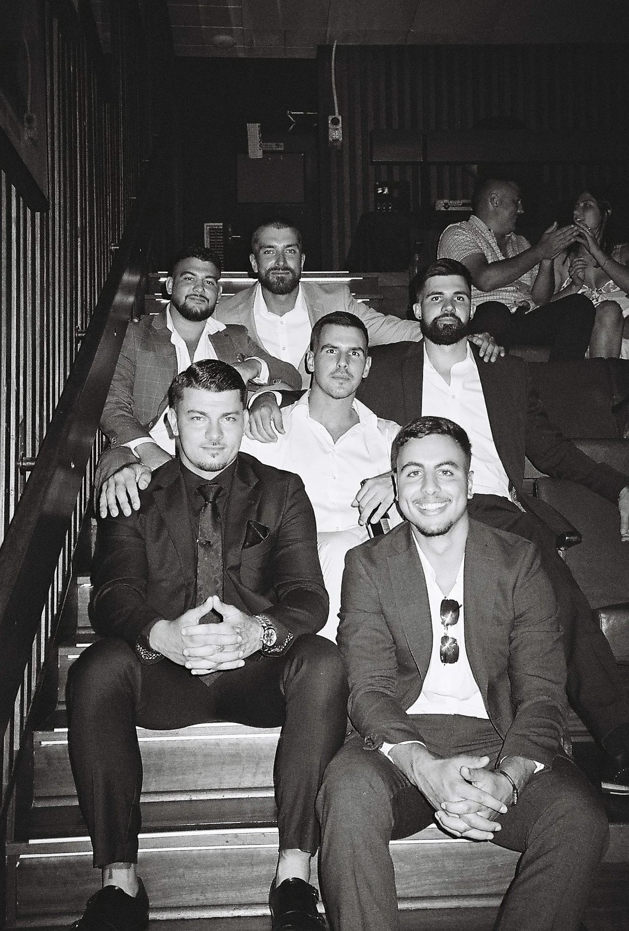 A group of six men sitting on a staircase at a social event, dressed in formal attire, with some smiling at the camera. In the background, two women are sitting together, engaging in conversation. The image is in black and white.