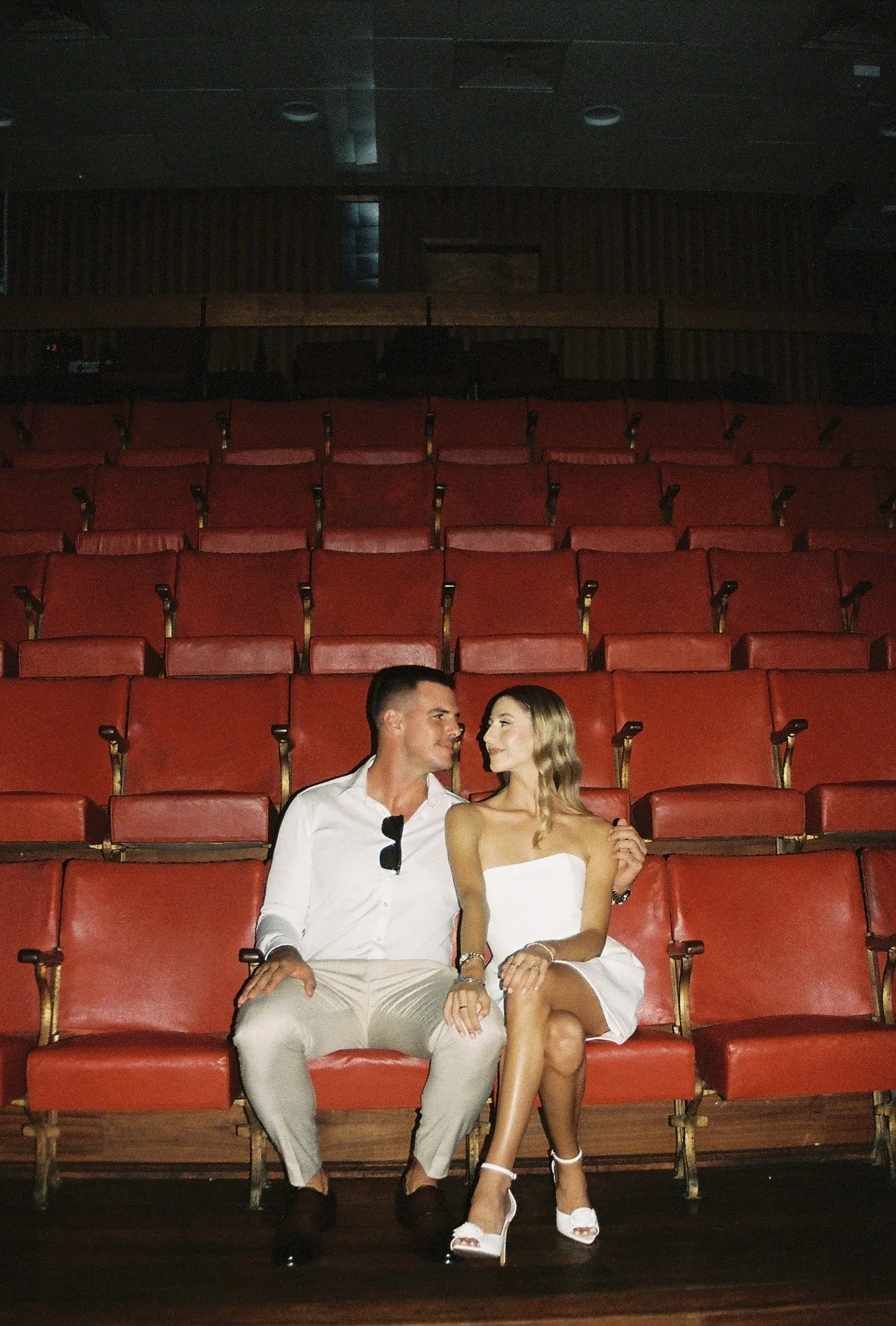 A young man and woman sitting together on red theater seats, looking at each other and smiling, in an empty auditorium.