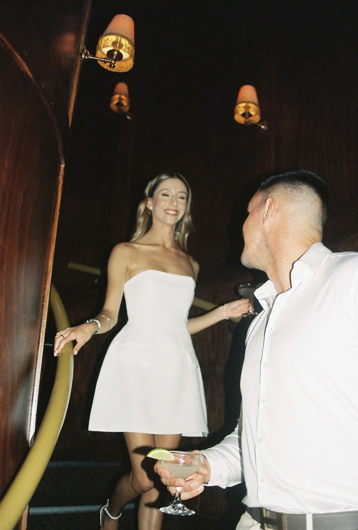 A woman in a white strapless dress smiling and standing on a staircase, looking at a man with a fade haircut and white shirt holding a cocktail with lime garnish, in a dimly lit room with dark wood walls and two wall-mounted lamps.