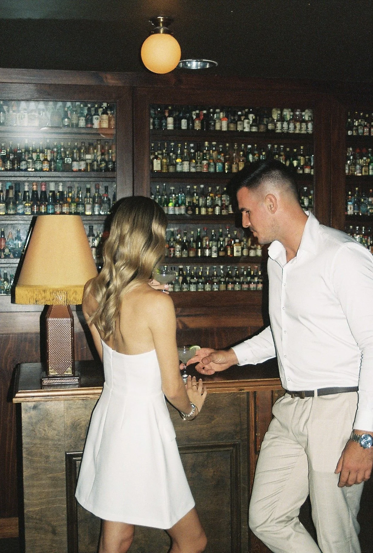 A man proposes to a woman in a bar with a large collection of liquor bottles behind them. The woman is wearing a white dress and has wavy, shoulder-length hair, while the man is dressed in a white shirt and light-colored pants.