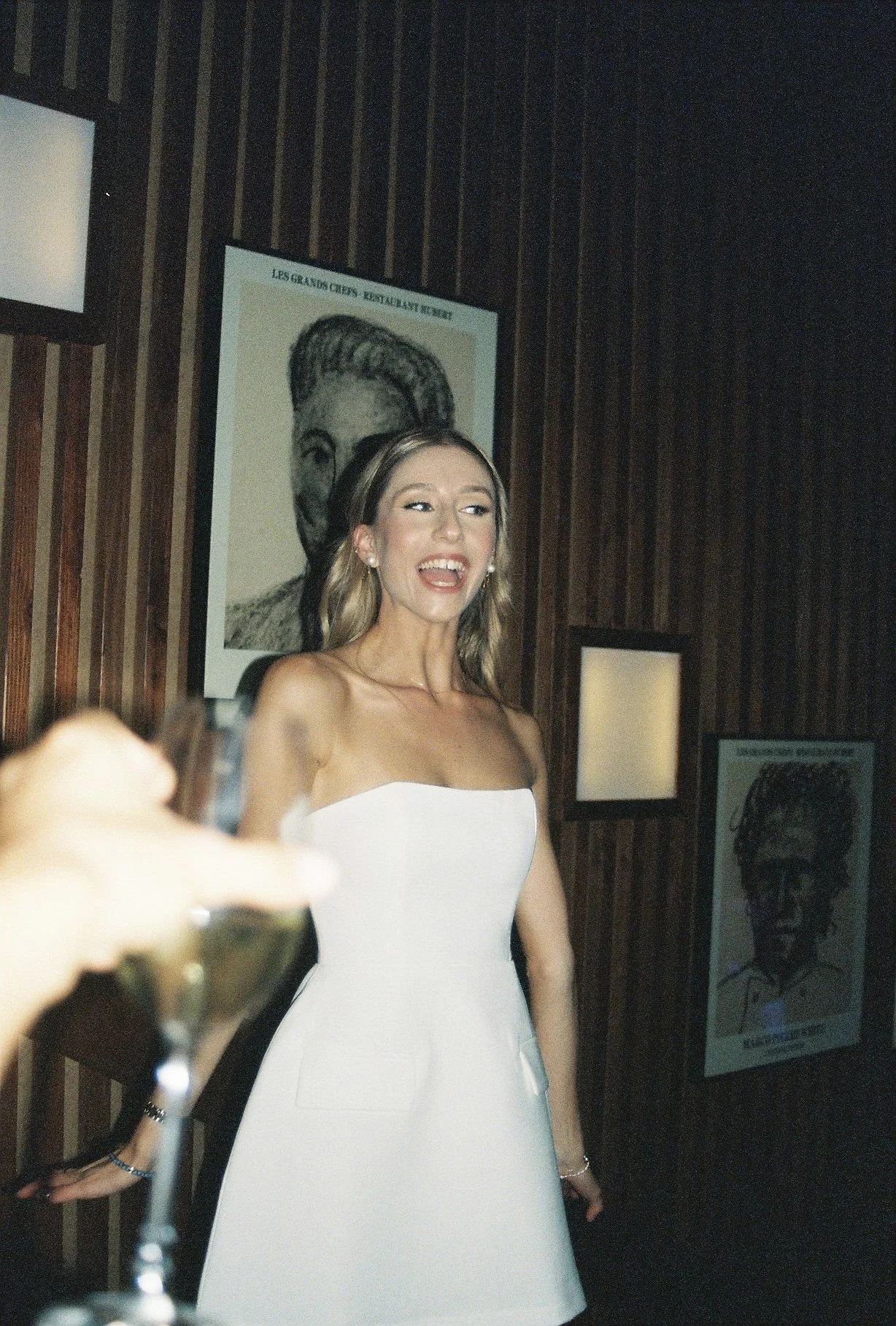 A woman in a strapless white dress standing in front of a wooden wall with framed artwork, smiling and appearing to be at a social event.
